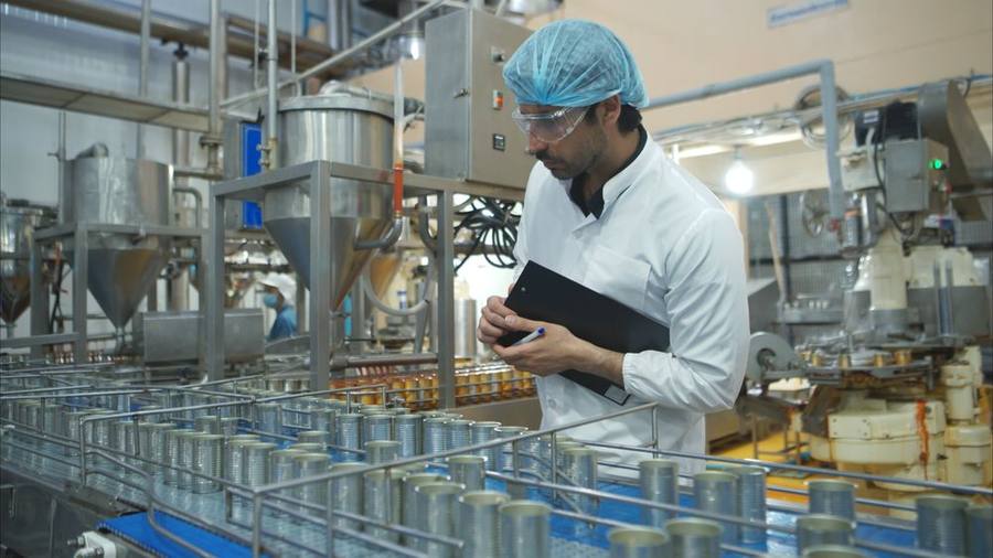 A quality officer in a lab coat, hairnet and safety glasses recording checks against product on a processing line