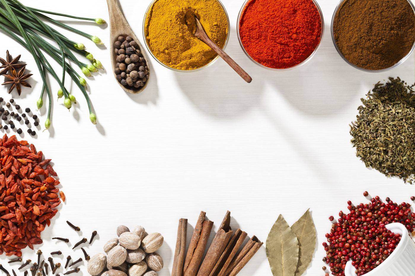 Turmeric, chilli and mixed ground spices in bowls, with star anise, peppercorns, cloves, nutmeg, cinnamon, bay leaves and dried herbs laid out around a clear centre